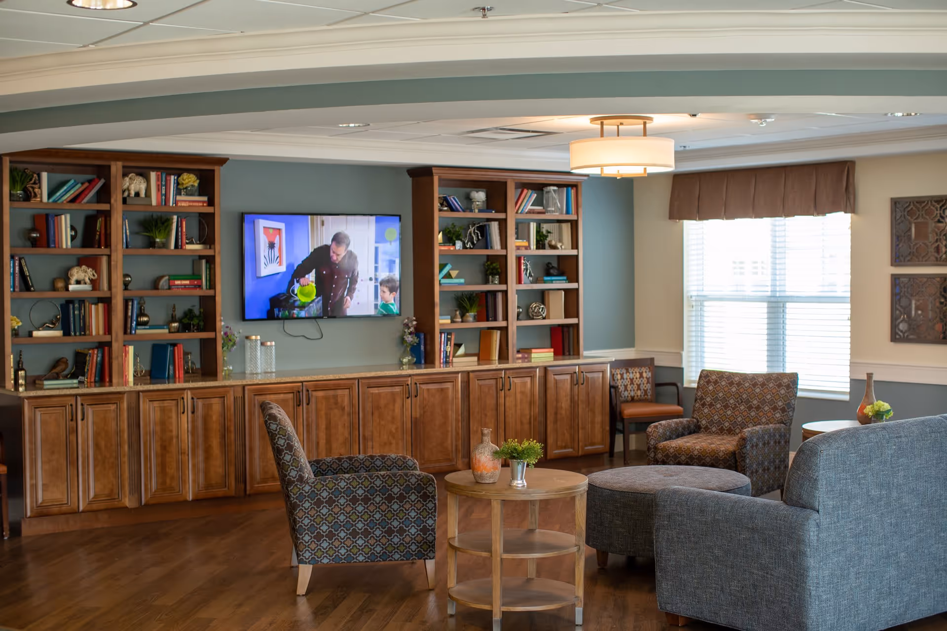 A cozy living room area with wooden bookshelves filled with books and decorative items, a wall-mounted TV showing a man and a child, patterned armchairs, a round wooden coffee table with small plants and a vase, a large window with blinds and a brown valance, and a ceiling light fixture.