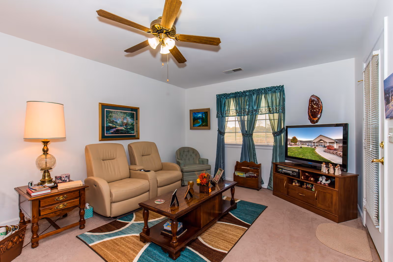 A cozy living room with two beige recliner chairs and a green armchair. There is a wooden coffee table with decorative items on a patterned rug. A wooden side table with a lamp and books is next to the recliners. A ceiling fan with lights is mounted on the white ceiling. The room has a window with teal curtains and a wooden TV stand with a flat-screen TV displaying an exterior view of a building. The walls are decorated with framed paintings and a wall clock.