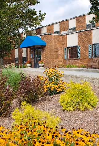 Exterior view of a brick building with a blue awning over the entrance. There are benches near the entrance and a landscaped garden with yellow and purple flowers in the foreground.