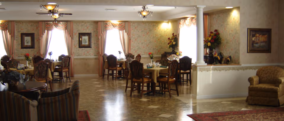 A warmly lit dining room with several tables covered in yellow tablecloths, each set with chairs and small floral centerpieces. The room features floral wallpaper, framed artwork on the walls, large windows with draped curtains, and a polished tile floor. There are also a few upholstered armchairs and decorative plants in the corner.