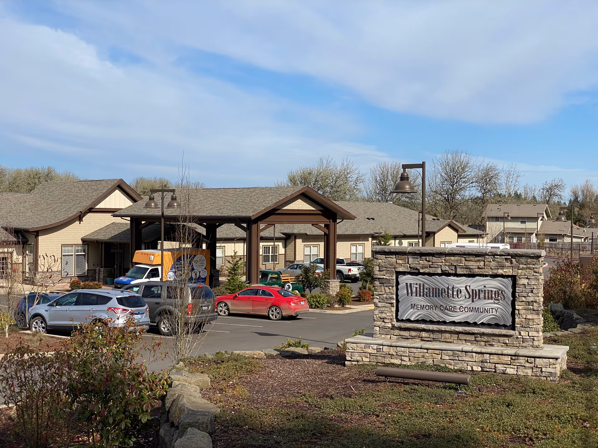 Exterior view of Willamette Springs Memory Care Community showing a stone sign with the facility name in front, a parking lot with several cars, and a building with a covered entrance under a partly cloudy sky.