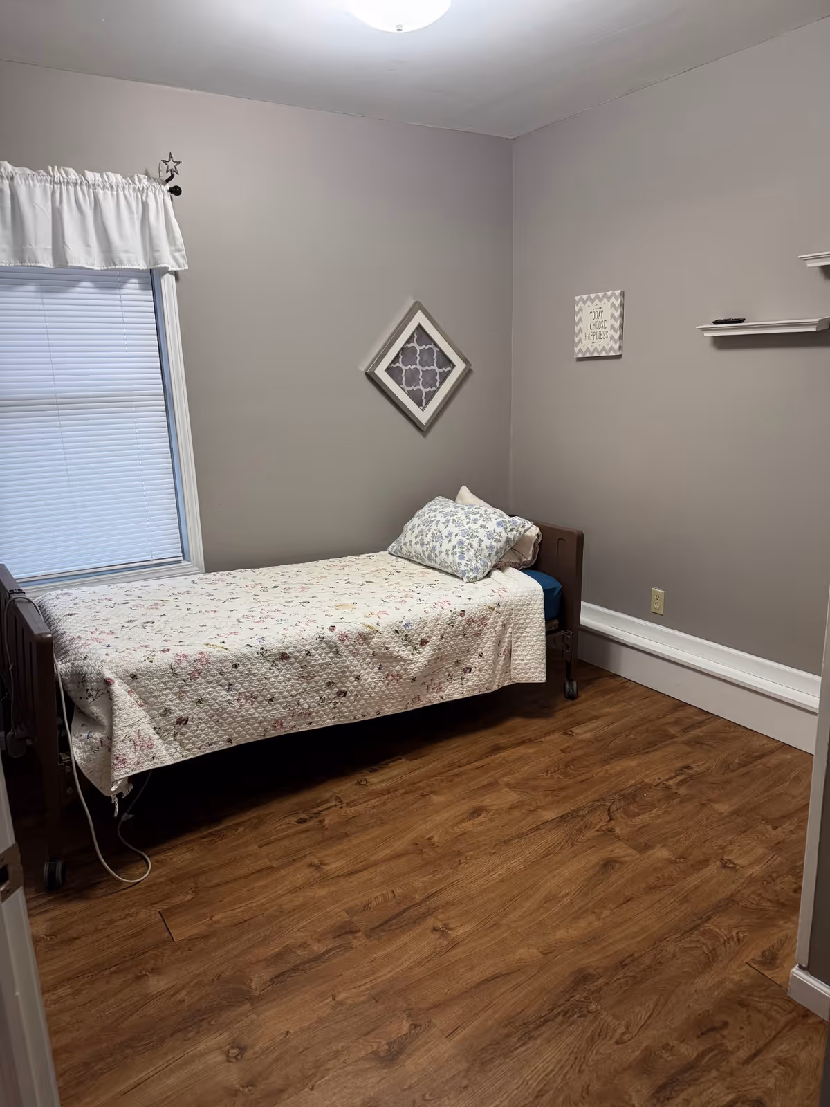 A small bedroom with a single bed covered with a floral quilt and a floral pillow. The room has light gray walls, wood flooring, a window with closed blinds and a white valance, a diamond-shaped wall decoration, a small framed sign on the wall, and two white floating shelves.