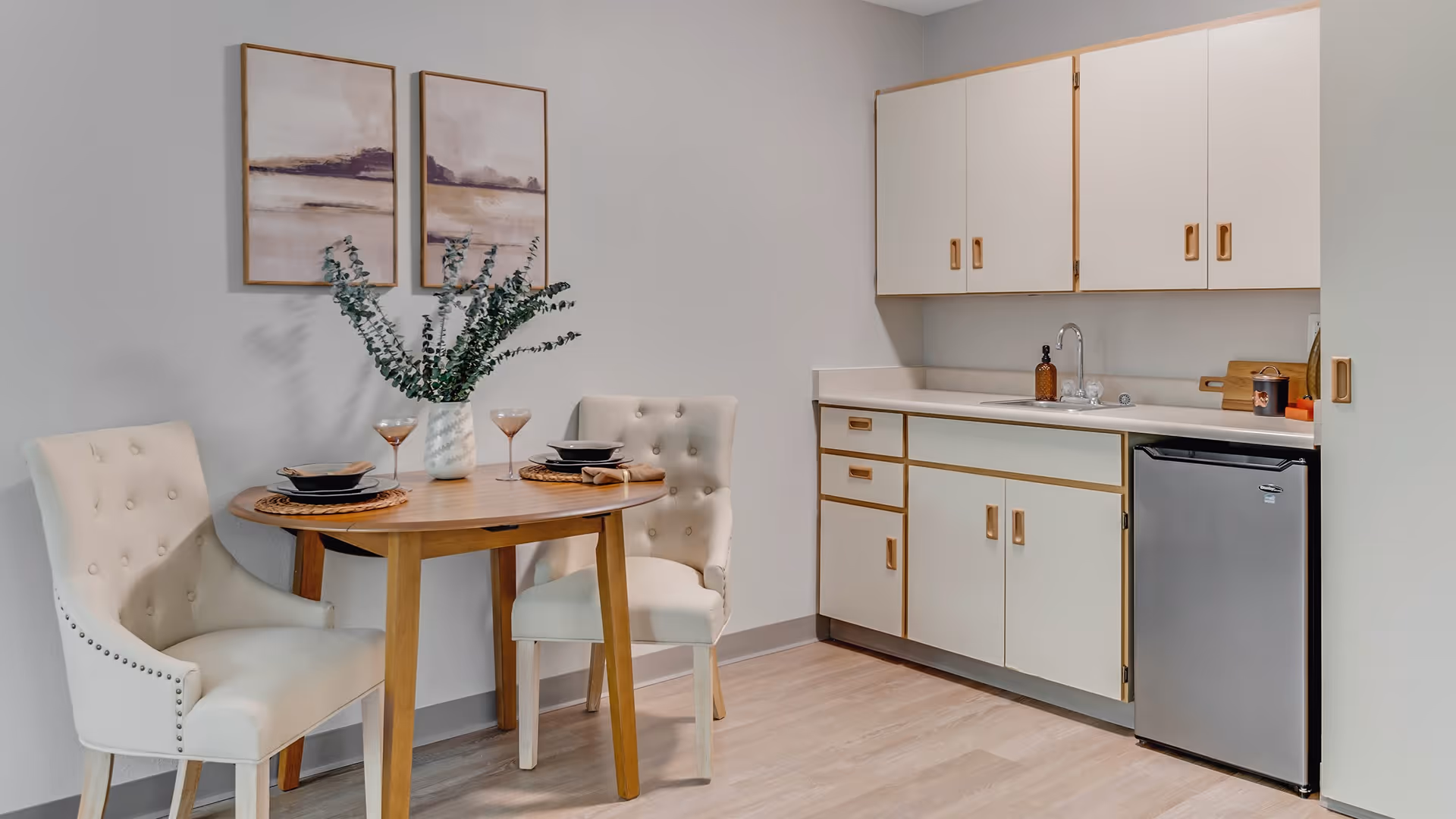 A small dining area with a round wooden table set for two with plates, bowls, and wine glasses. Two upholstered cream-colored chairs with button tufting are placed around the table. Behind the dining area is a kitchenette with white cabinets, a small stainless steel refrigerator, a sink, and a soap dispenser. Two framed abstract art pieces hang on the light gray wall above the table.