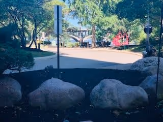 Outdoor area with large rocks in the foreground, a paved pathway, trees providing shade, and playground equipment visible in the background under a clear sky.