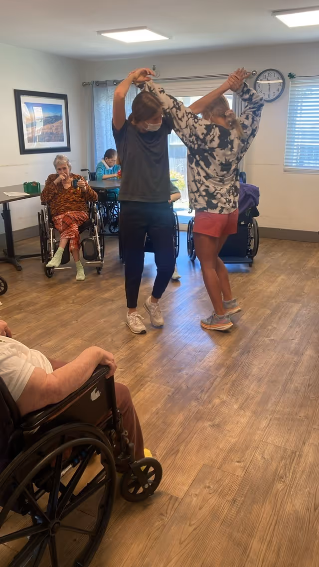 Two women dancing together in a room with wooden flooring, while several elderly individuals in wheelchairs watch. The room has white walls, a clock, a window with blinds, and a framed picture on the wall.