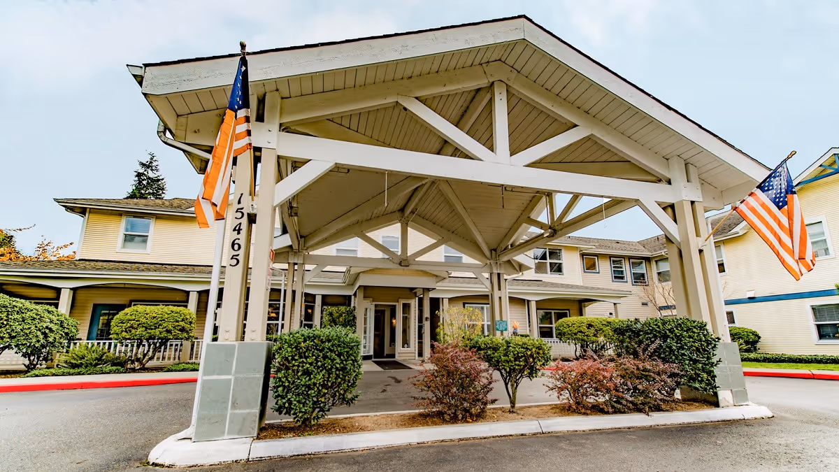 Covered main entrance canopy of a multi-story senior living building with American flags and landscaped shrubs.