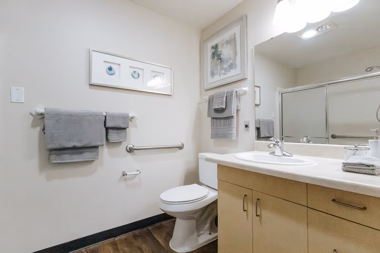 A clean and modern bathroom featuring a toilet, a sink with a countertop and cabinet, a large mirror, and a shower with sliding glass doors. The walls are decorated with framed artwork and gray towels hang on towel racks.