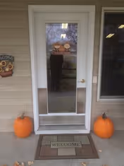 A white door with a glass panel in the center, flanked by two orange pumpkins on the concrete porch. A welcome mat with the word 'WELCOME' is placed in front of the door. The door has a small decorative sign with pumpkins and the word 'Welcome' on it. The exterior wall is beige with horizontal siding, and there is a window to the right of the door.