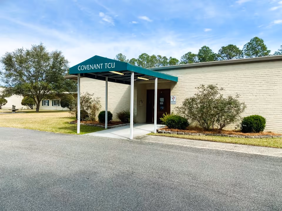Exterior view of a single-story building with a green canopy labeled 'COVENANT TCU' over the entrance. The building is light-colored brick with bushes and small trees planted along the front. There is a paved driveway or parking area in front and a grassy area with more trees in the background under a partly cloudy sky.