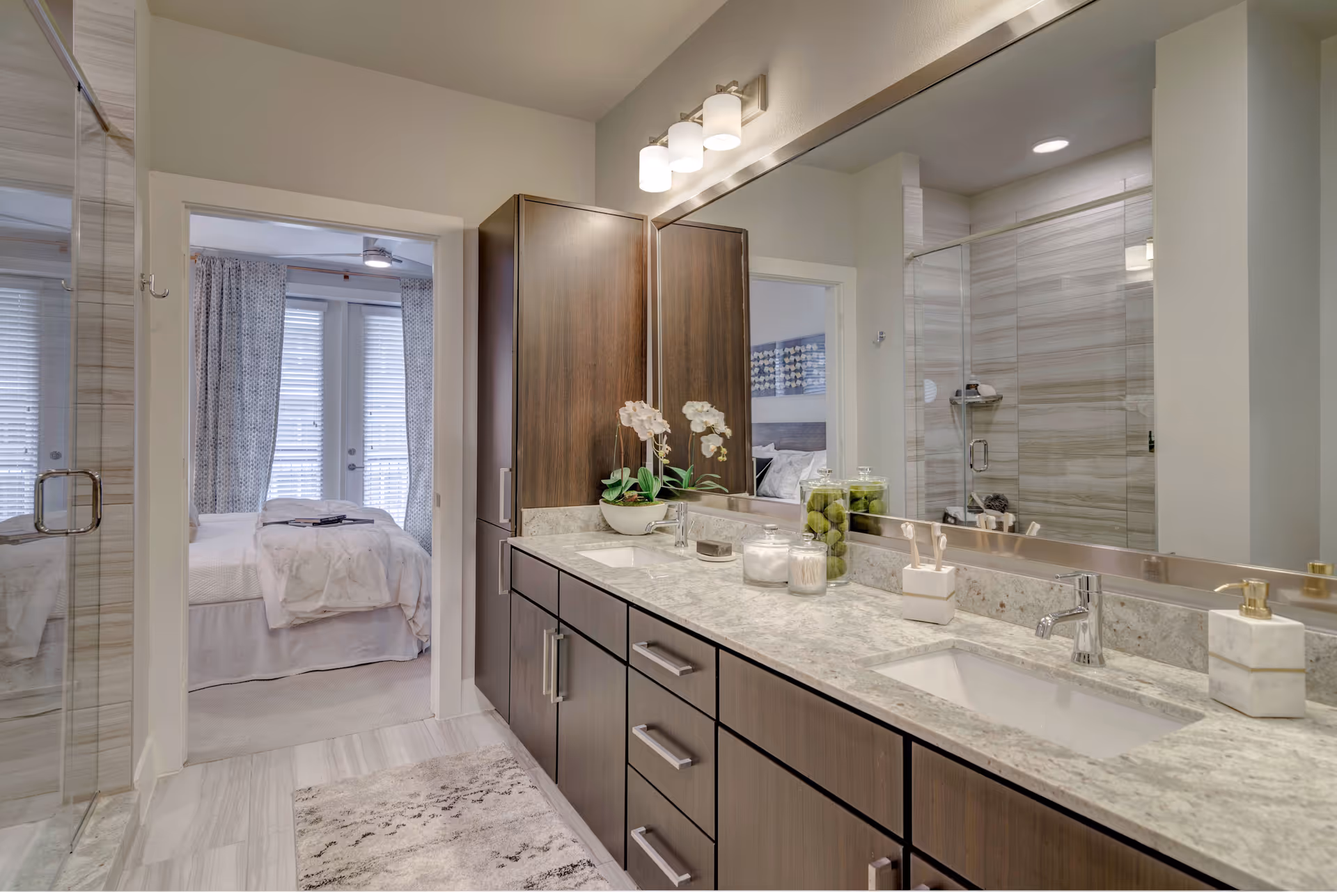Modern bathroom with a double vanity and large mirror, glass-enclosed shower, and a view into a bedroom.
