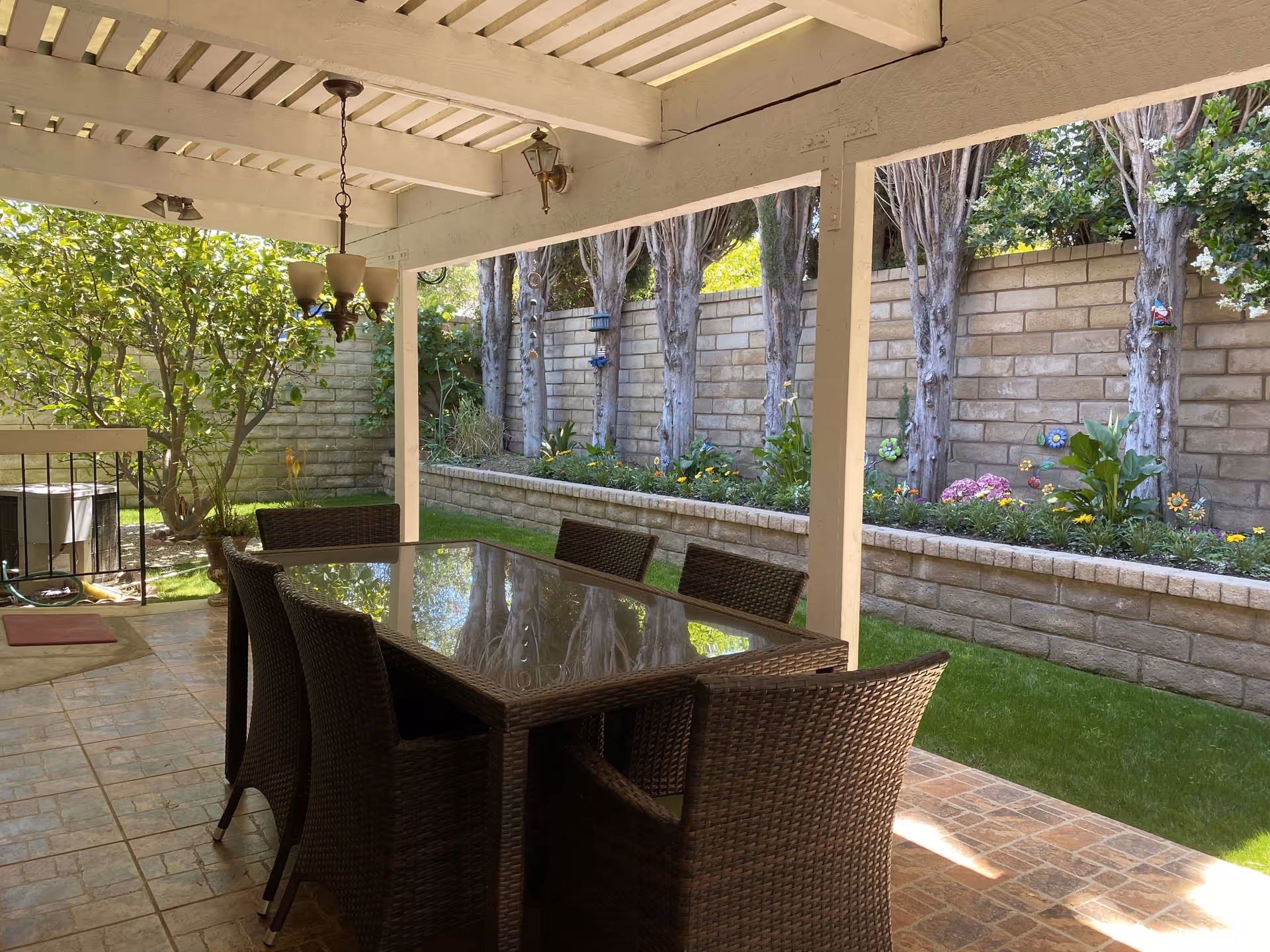 Covered patio with a glass-topped wicker dining table and chairs overlooking a landscaped yard with a low brick retaining wall and trees.