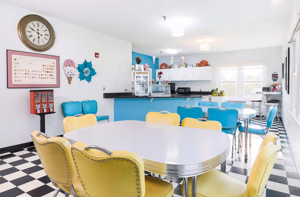 Bright and cheerful dining area with retro-style yellow and turquoise chairs around white tables. The room features a black and white checkered floor, a blue counter with a popcorn machine, white cabinets, and wall decorations including a large clock and ice cream themed art.
