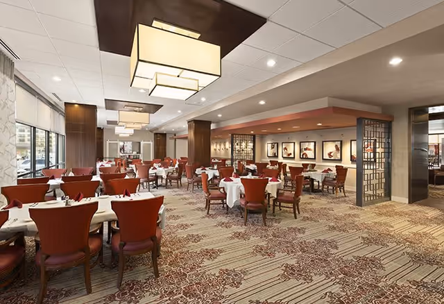 A spacious dining room with multiple tables covered in white tablecloths and set with red napkins. The room features red upholstered chairs, large rectangular ceiling lights, patterned carpet, and framed artwork on the walls. Large windows allow natural light to enter the space.