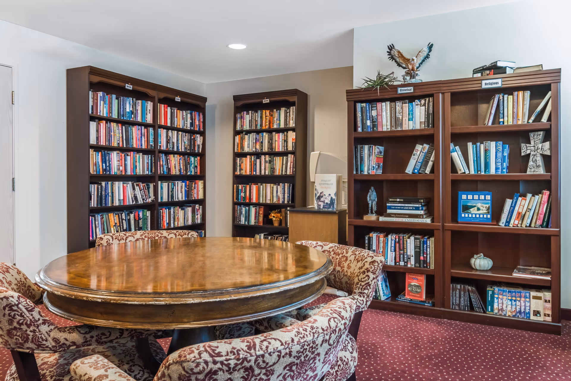 A cozy reading room with three large wooden bookshelves filled with books. In front of the bookshelves is a round wooden table surrounded by four upholstered chairs with a floral pattern. The room has a carpeted floor with a red and white dotted pattern and white walls.