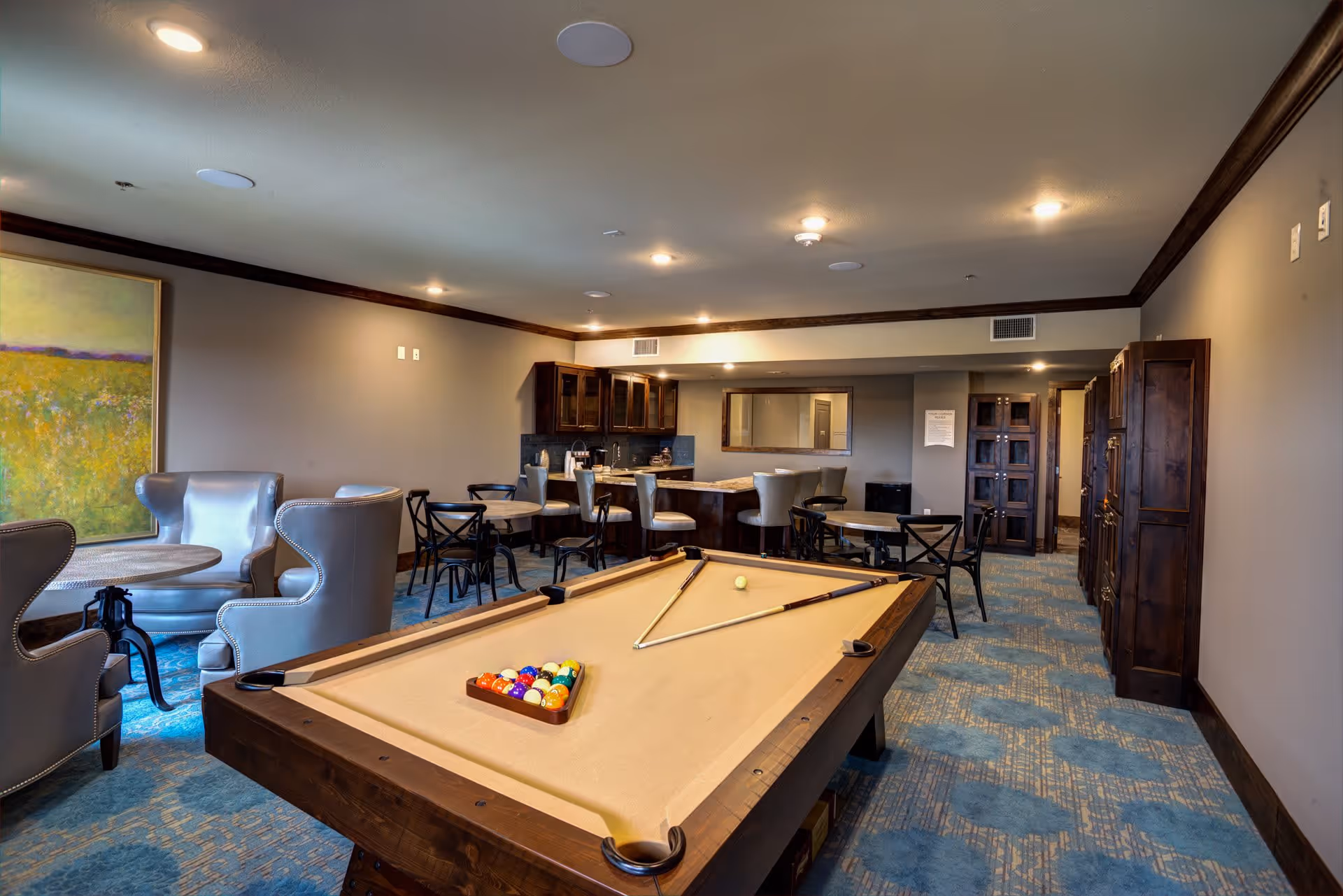 A recreational room featuring a pool table with balls and cues arranged on it, surrounded by several seating areas including armchairs, small round tables with chairs, and a bar counter with stools. The room has a blue patterned carpet, beige walls with dark wood trim, and a large painting on one wall.