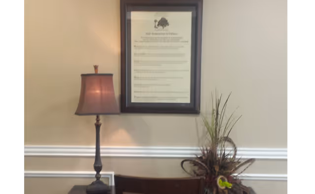 A framed statement of values hanging on a beige wall above a wooden chair. To the left of the chair is a table lamp with a dark base and a brown lampshade. To the right is a decorative arrangement of tall grasses and flowers.