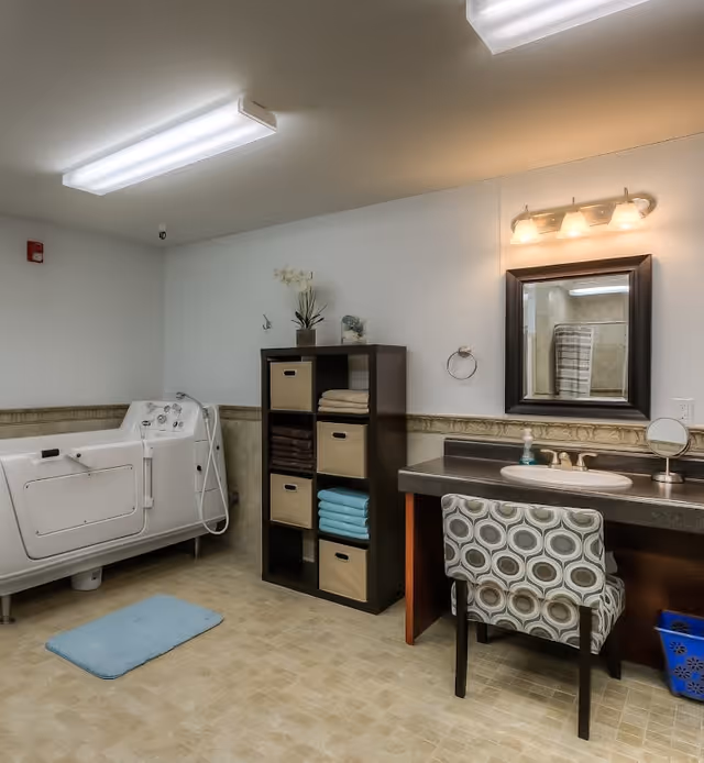 A bathroom with a walk-in bathtub on the left, a shelving unit with towels and storage bins in the center, and a vanity with a sink, mirror, and patterned chair on the right. The room has tiled flooring and bright overhead lighting.