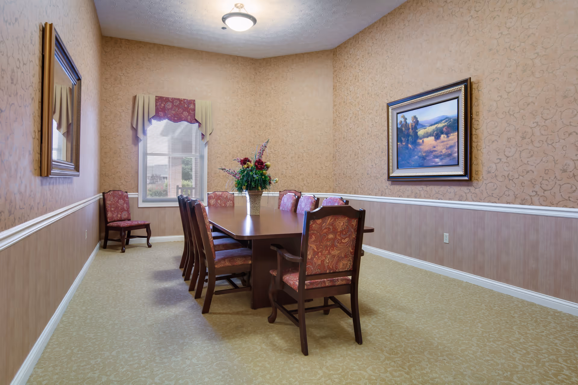 A formal dining room with a long wooden table surrounded by eight upholstered chairs with a floral pattern. A floral centerpiece is placed on the table. The room has beige patterned wallpaper with a white chair rail, a window with valance curtains, a framed landscape painting on the wall, and a ceiling light fixture.