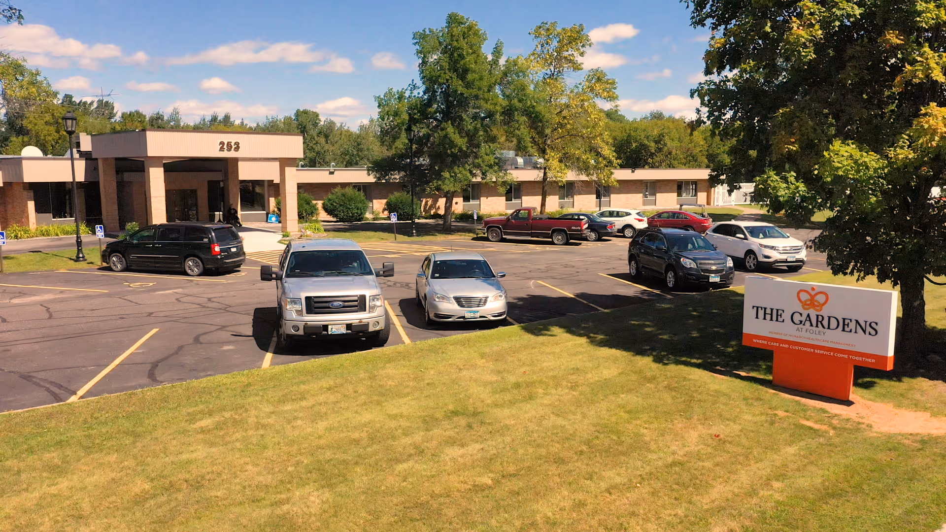 Exterior view of The Gardens at Foley senior living facility showing a parking lot with several parked cars, a building entrance with the number 253 above it, and a sign with the facility's name on a grassy area under a partly cloudy sky.