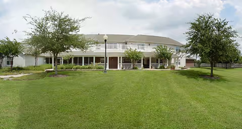 Front view of a two-story building with a covered porch and upper balcony, a wide green lawn, and several trees under a partly cloudy sky.