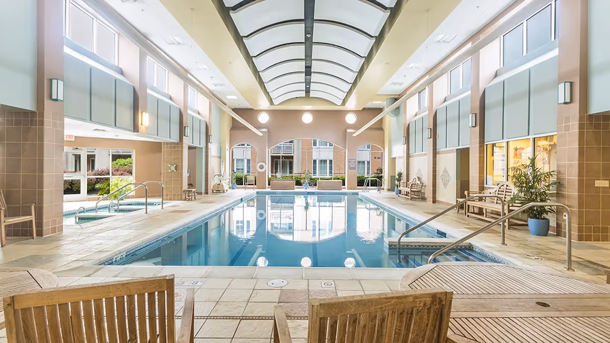 Indoor swimming pool area with a high arched ceiling and large windows letting in natural light. The pool is surrounded by tiled flooring and wooden benches, with handrails for easy access. There are potted plants and a hot tub visible on the left side.