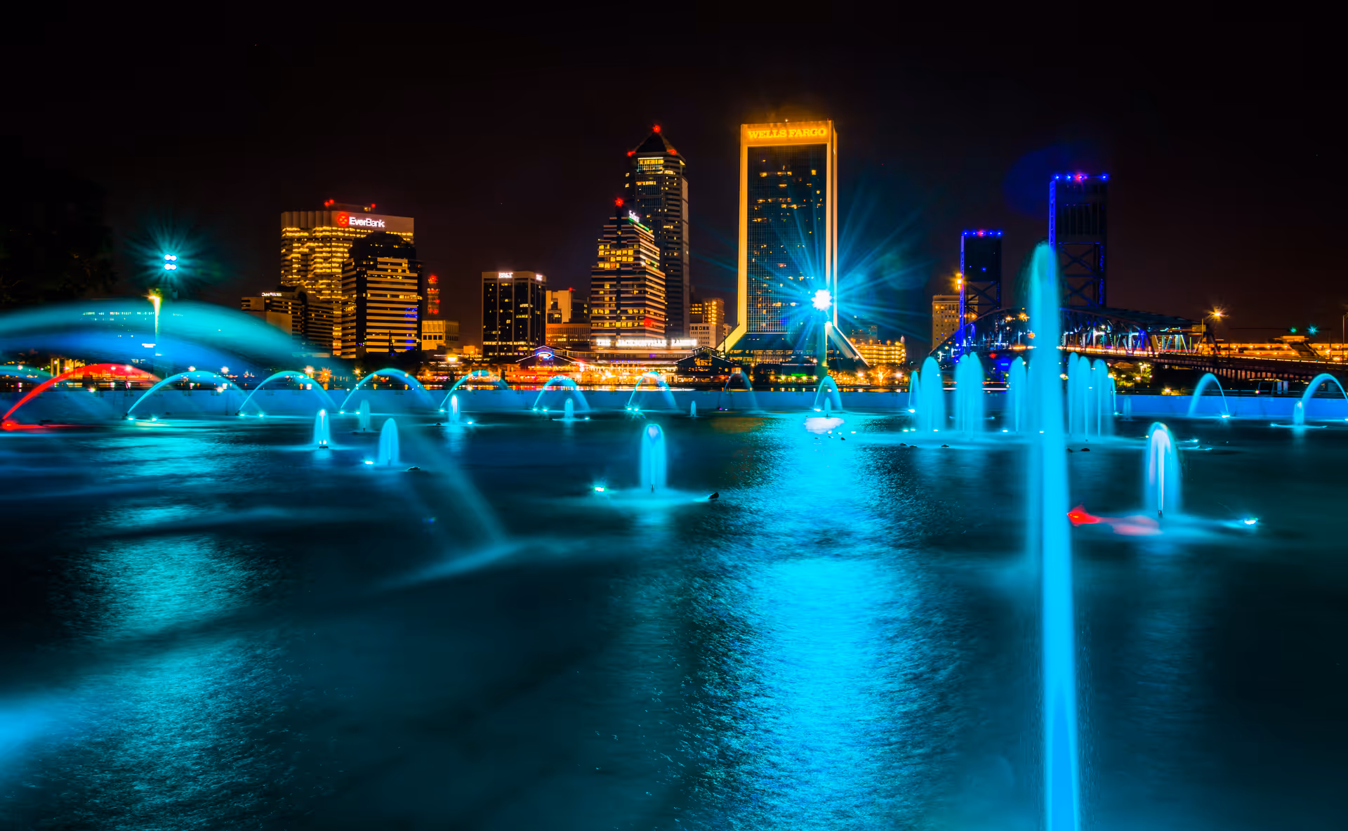 Nighttime cityscape featuring illuminated buildings and a bridge in the background, with a large fountain in the foreground shooting streams of water lit by blue lights.
