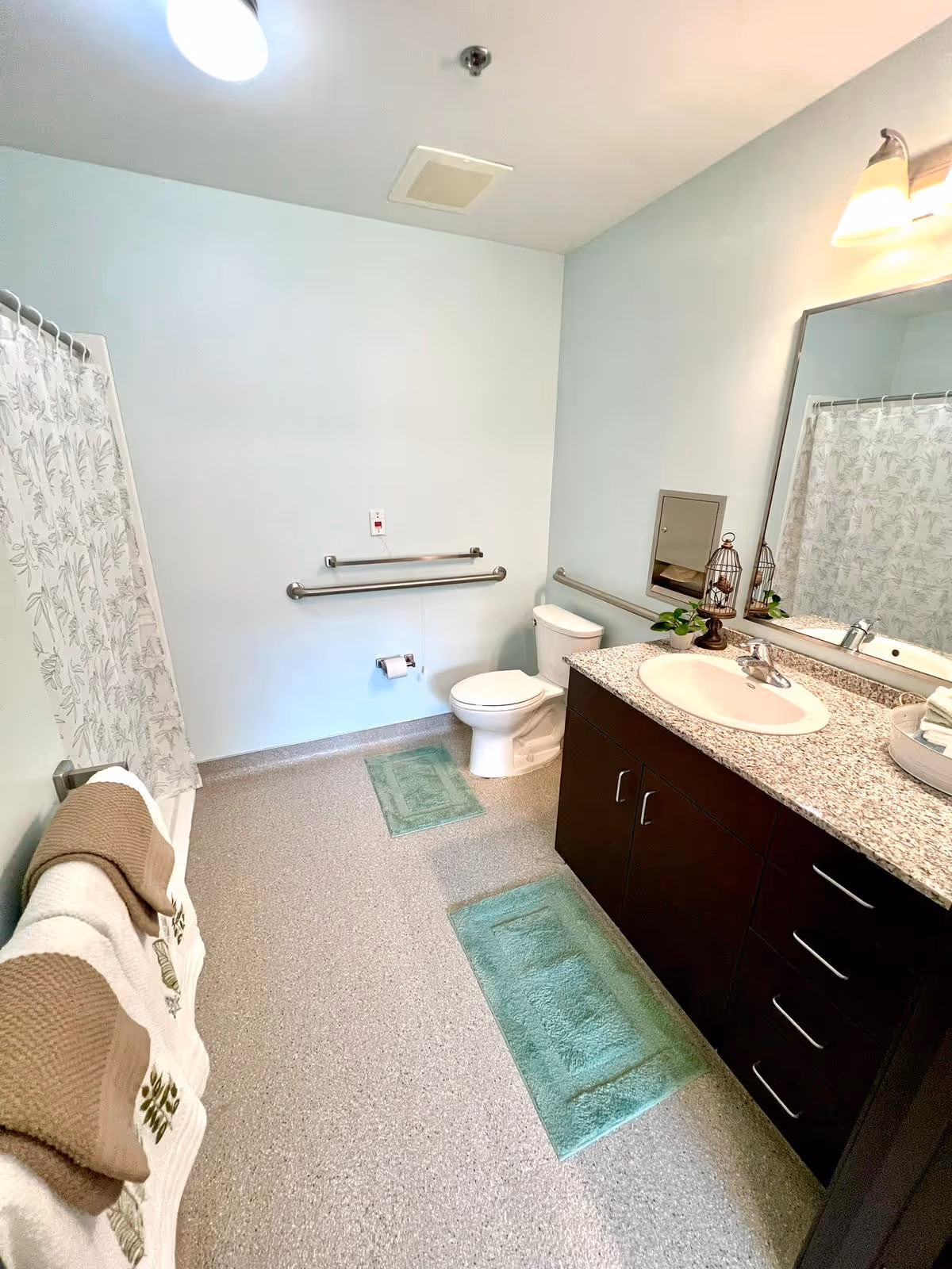 A clean and accessible bathroom with a toilet, a sink with a granite countertop, a large mirror, and a shower with a floral-patterned curtain. There are two green bath mats on the floor, grab bars near the toilet, and neatly folded towels on a rack.