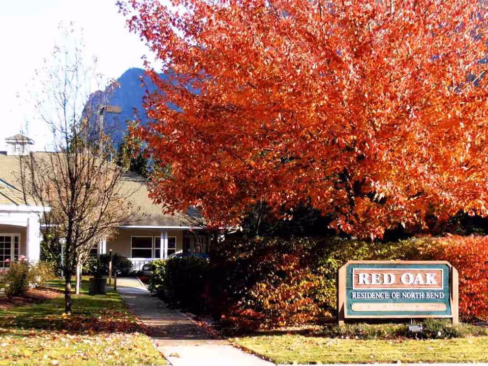 Exterior view of Red Oak Residence senior living facility with a large tree displaying vibrant red autumn leaves and a sign that reads 'RED OAK RESIDENCE OF NORTH BEND' near the sidewalk leading to the building entrance.