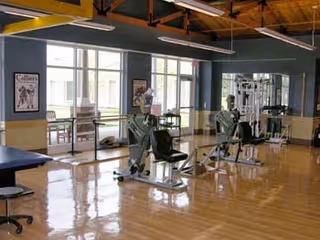 Interior view of a fitness room with exercise equipment including stationary bikes and weight machines. The room has large windows letting in natural light, wooden floors, and exposed ceiling beams. There are framed pictures on the walls and a bench visible on the left side.