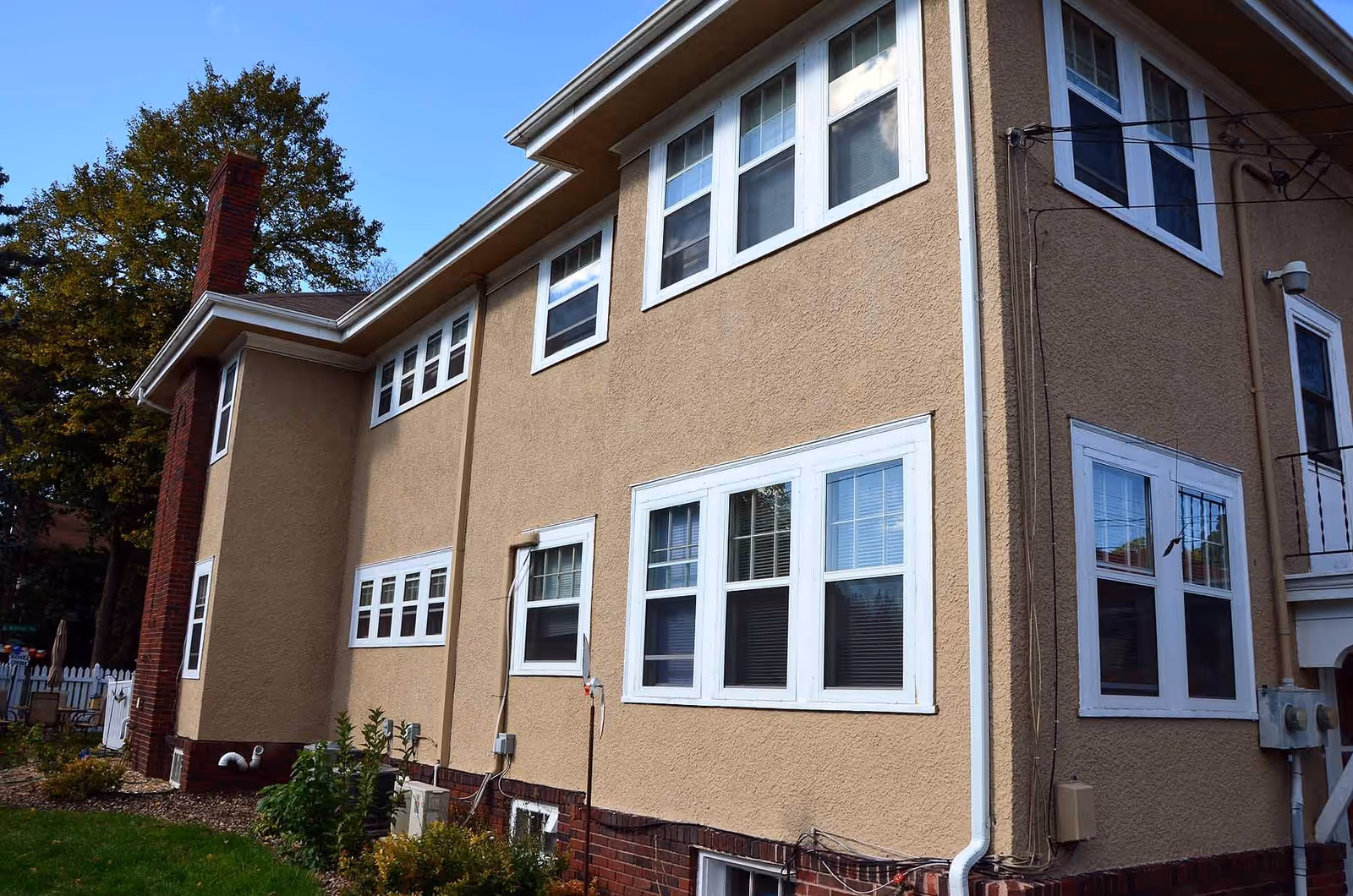 Exterior view of a two-story beige building with multiple white-framed windows and a red brick chimney, surrounded by some greenery and a tree in the background under a clear blue sky.