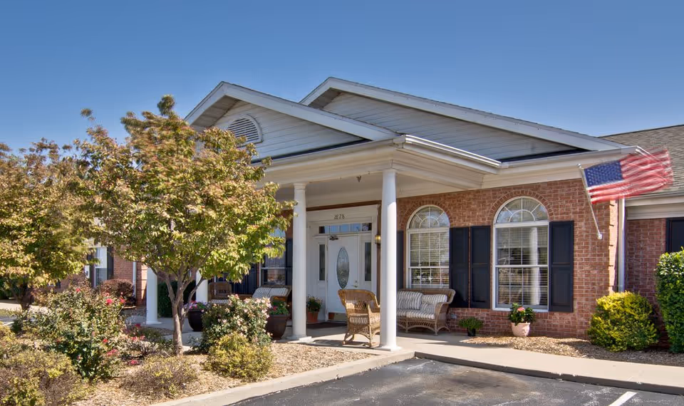 Exterior view of Spring Ridge Assisted Living facility showing a brick building with white columns at the entrance, a porch with wicker chairs and a bench, an American flag mounted on the right side, and landscaped bushes and trees in front under a clear blue sky.