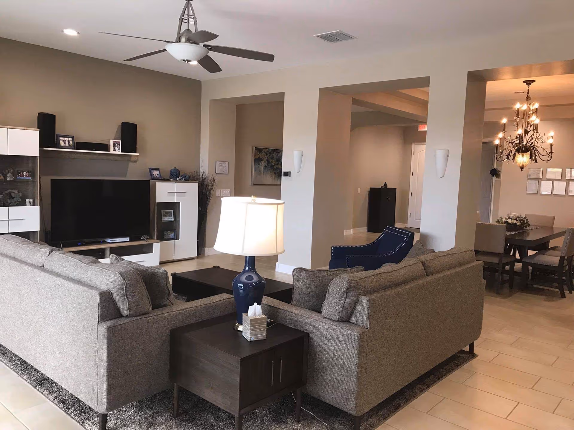 A modern living room with two gray sofas facing a flat-screen TV mounted on a white entertainment unit. A dark wooden side table with a blue lamp and tissue box is placed between the sofas. In the background, there is a dining area with a chandelier hanging above a dining table surrounded by chairs. The room has beige walls, tiled floors, and a ceiling fan with lights.