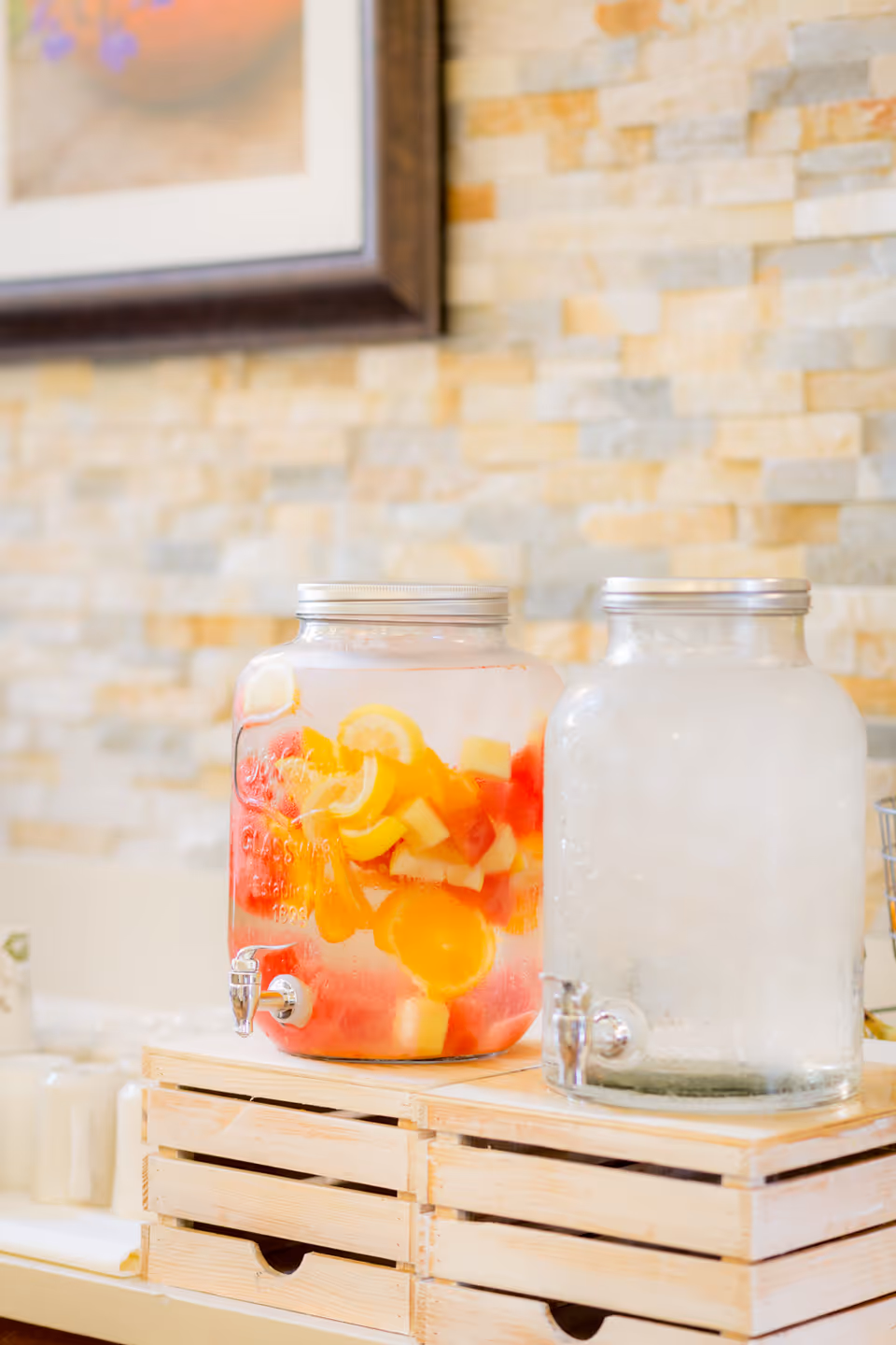 Two large glass beverage dispensers on a wooden crate stand. One dispenser contains water infused with slices of lemon, orange, and other fruits, while the other is filled with ice water. The background features a light-colored stone wall and a partially visible framed picture.