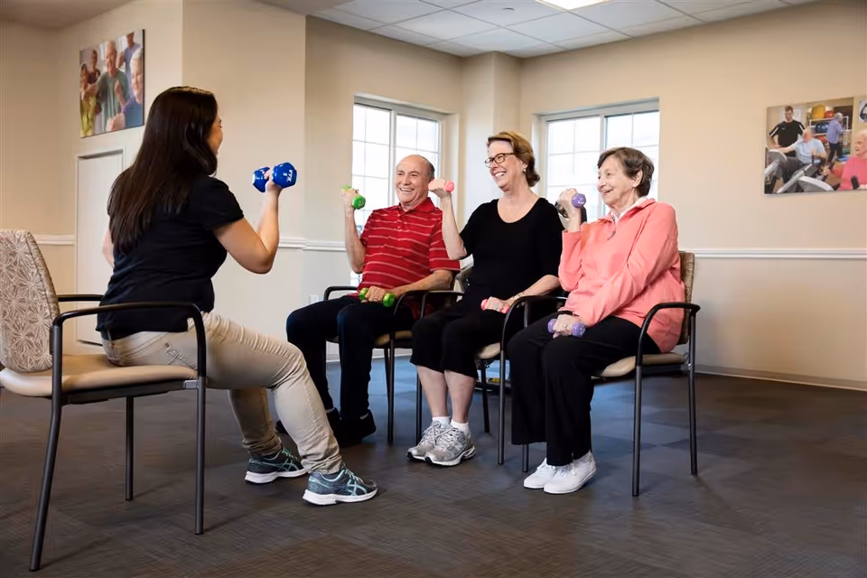 A group of three elderly people sitting on chairs in a room, each holding small dumbbells and participating in a seated exercise session led by a younger woman also holding dumbbells.