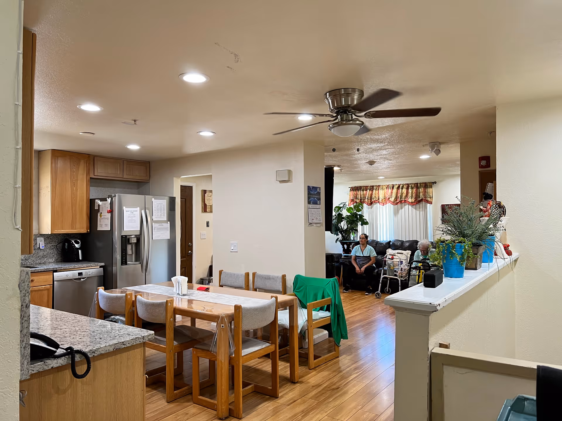 Open-plan kitchen and dining area with a wooden table and chairs, refrigerator and countertops, opening into a living room where two people sit.