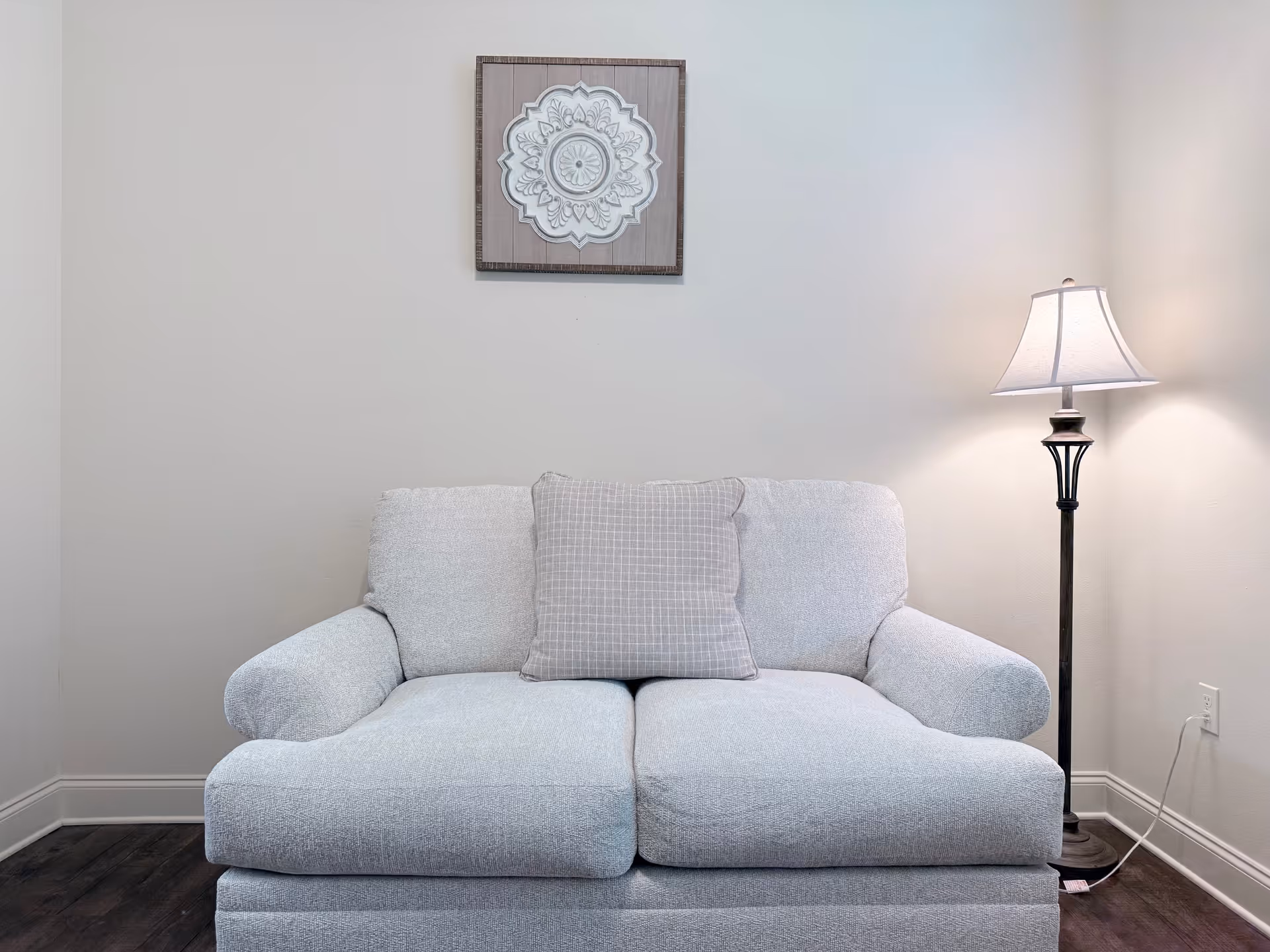 A cozy living room corner featuring a light gray loveseat with a single checkered cushion in the center. To the right of the loveseat is a tall floor lamp with a white lampshade, casting a warm light on the beige wall behind. Above the loveseat hangs a framed decorative artwork with a floral mandala design. The floor is dark wood, and there is a white electrical outlet with a plugged-in cord visible near the lamp.