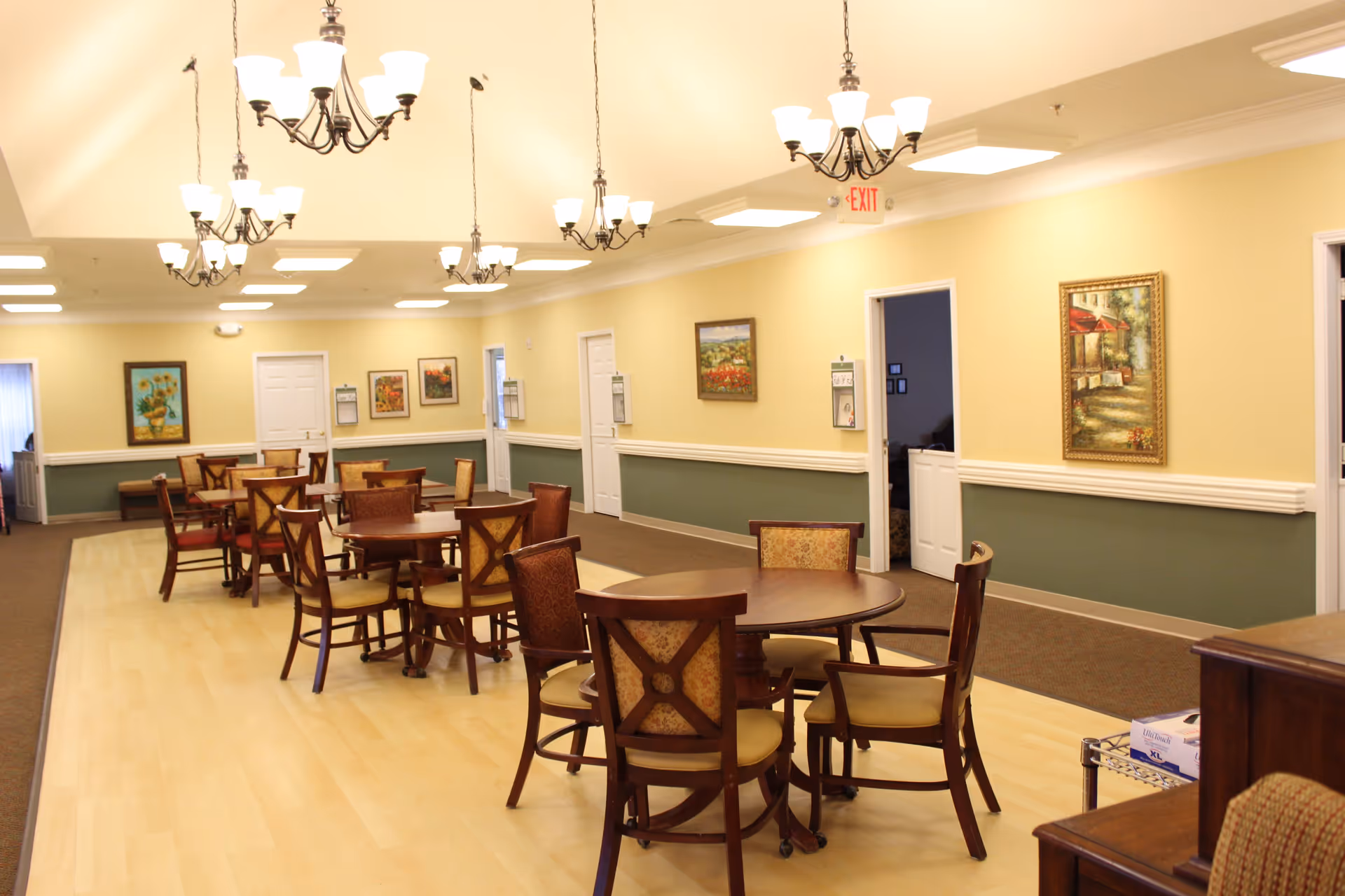 A spacious dining room with multiple round wooden tables and cushioned chairs arranged neatly. The room has light yellow walls with green wainscoting, several framed paintings, and multiple chandeliers hanging from the ceiling providing warm lighting. There are several doors along the walls and an exit sign above one door.