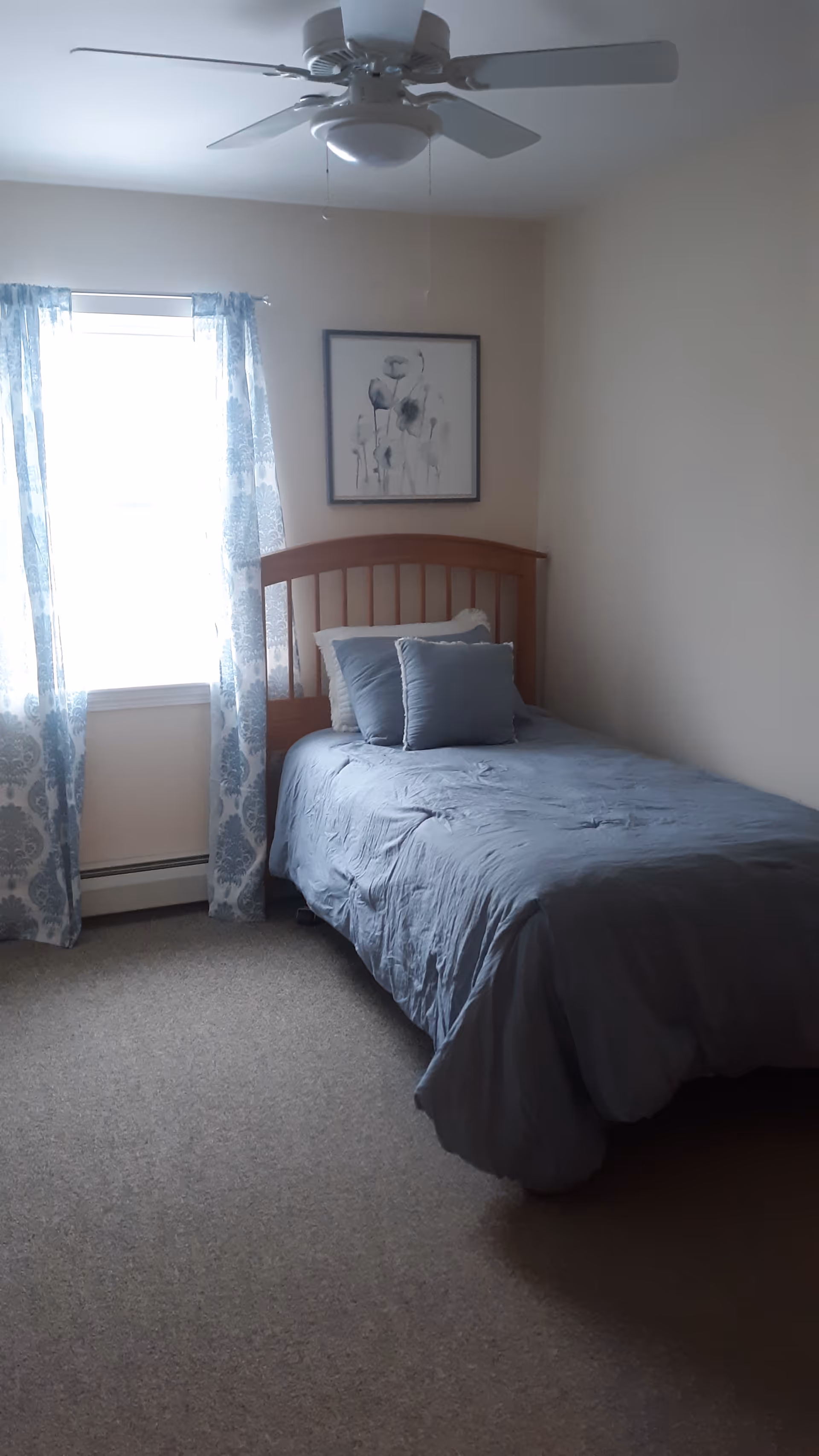 A small bedroom with a single bed covered in a blue comforter and two blue pillows. The bed has a wooden headboard. There is a window with blue patterned curtains to the left of the bed, and a framed floral artwork hangs on the wall above the headboard. A white ceiling fan is mounted on the ceiling.