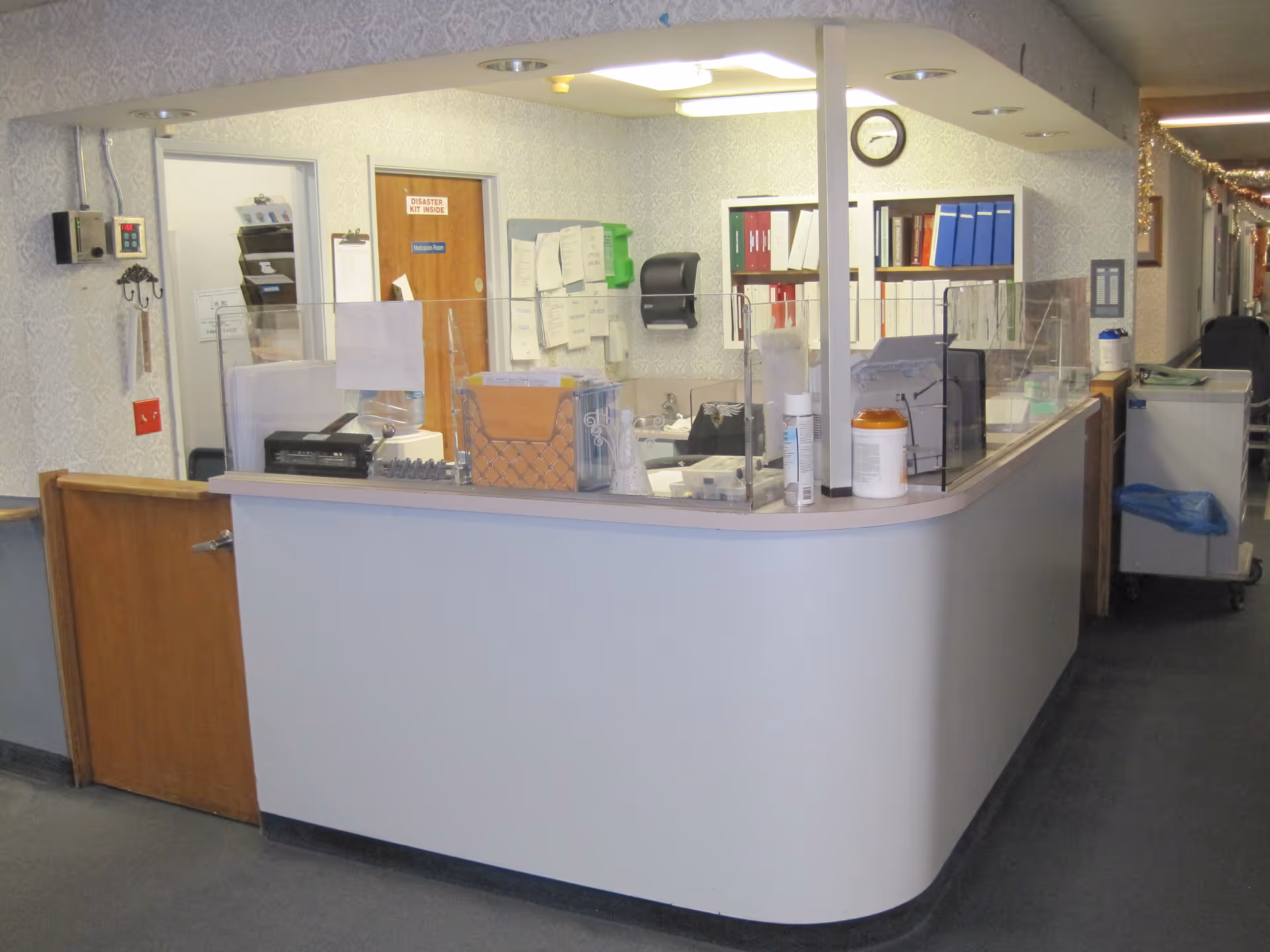 Reception desk area inside a facility with a curved counter, clear protective barriers, office supplies, and a clock on the wall. There are doors and shelves with binders in the background, and a hallway decorated with garlands is visible to the right.
