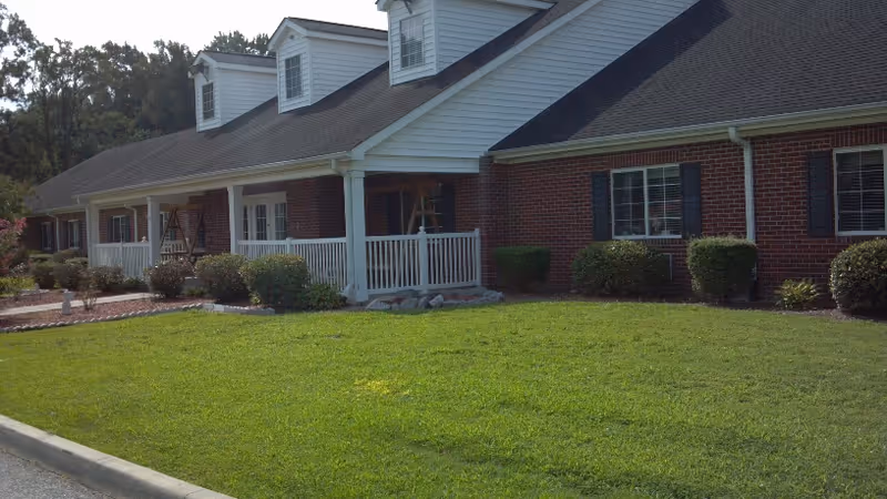 Exterior view of a single-story brick building with white trim and a covered porch with white railings. The building is surrounded by a well-maintained green lawn and some bushes. The sky is clear and trees are visible in the background.