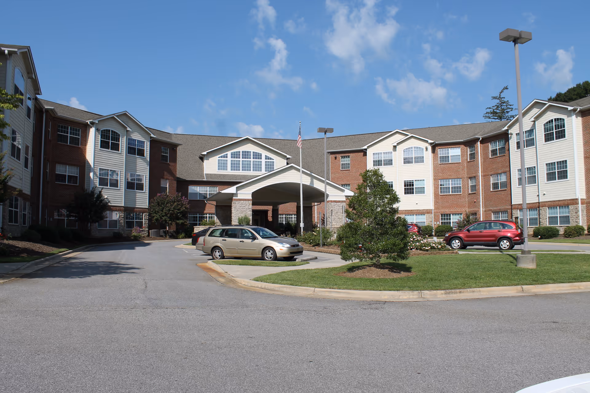 Front exterior of a three-story senior living building with a covered entrance, parked cars, and a landscaped driveway under a blue sky.