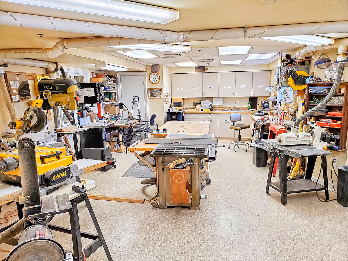 Interior woodworking workshop with multiple power tools, workbenches, and storage cabinets.