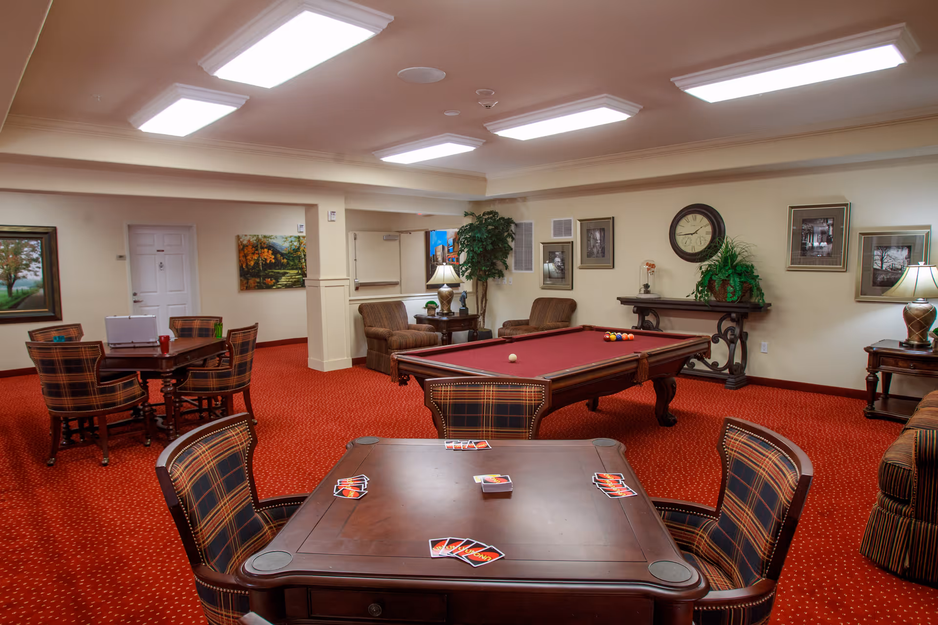 A cozy game room with red carpet and beige walls featuring a pool table with balls set up, a card table with cards laid out, several plaid upholstered chairs, two armchairs near a side table with a lamp, framed artwork on the walls, a clock above a decorative console table with a plant, and additional tables and chairs in the background.