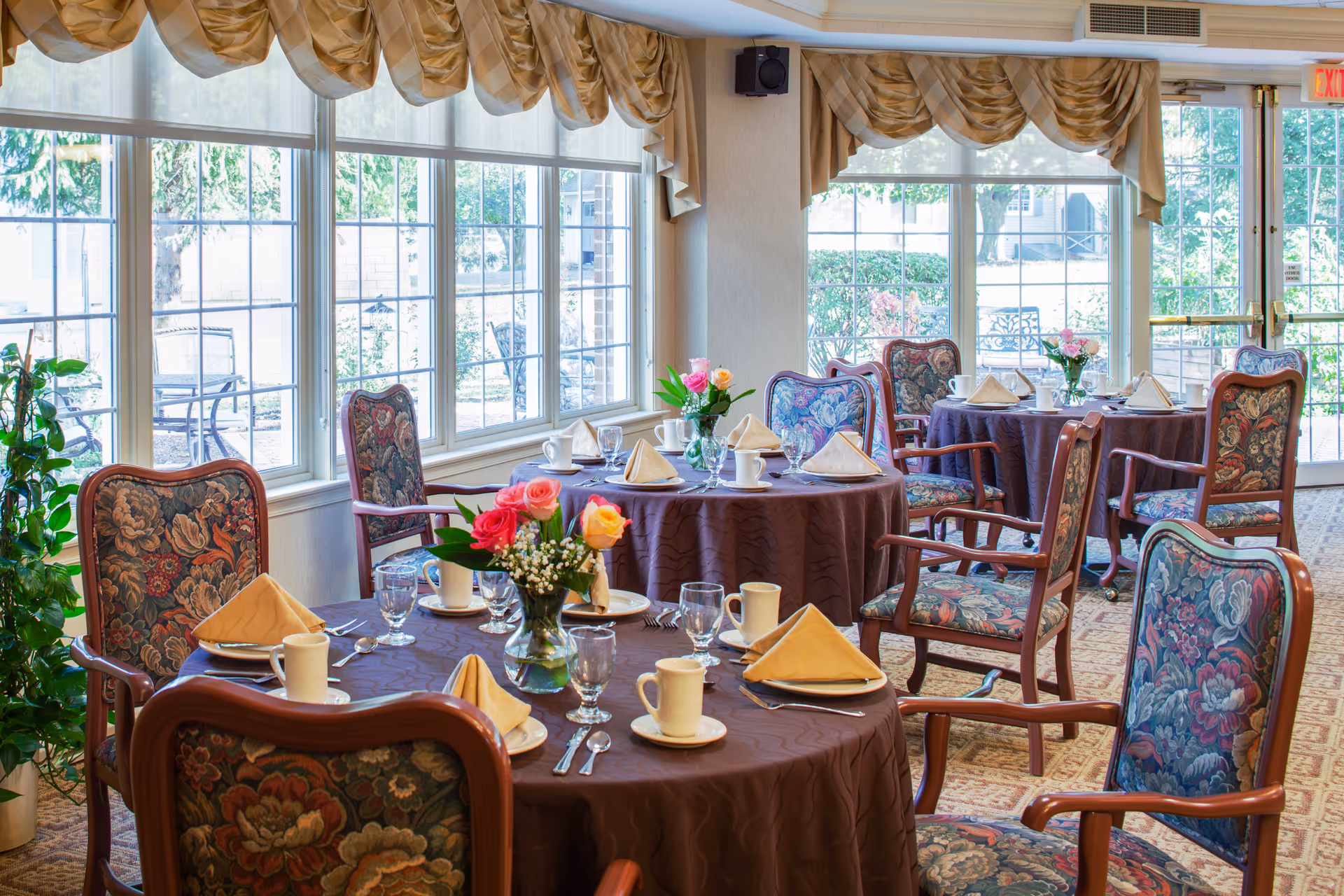 A bright dining room with round tables covered in brown tablecloths, each set with beige folded napkins, cups, glasses, and silverware. Floral upholstered wooden chairs surround the tables. Large windows with beige valances let in natural light and offer a view of outdoor greenery. Vases with colorful flowers decorate the tables.