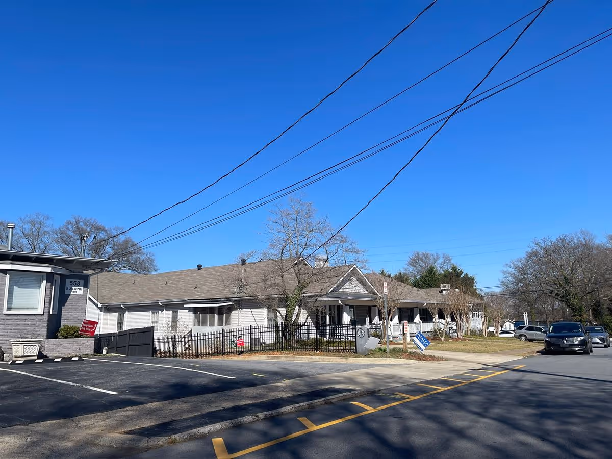 Front exterior of a single-story assisted living facility with a fenced yard, parking area, and clear blue sky.