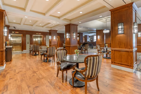 A spacious interior common area with wooden floors and wood-paneled columns. Several round tables with striped upholstered chairs are arranged throughout the room. The ceiling has recessed lighting and decorative beams. Two elevators with stainless steel doors are visible in the background.
