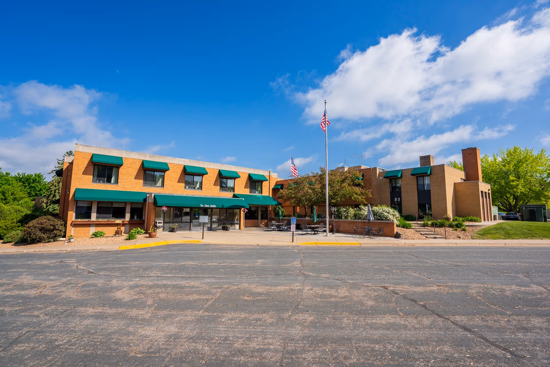 Exterior view of The Glenn Buffalo facility, a two-story brick building with green awnings over the windows and entrance. There are two American flags on flagpoles in front of the building, outdoor seating with tables and umbrellas, and surrounding greenery under a blue sky with some clouds.