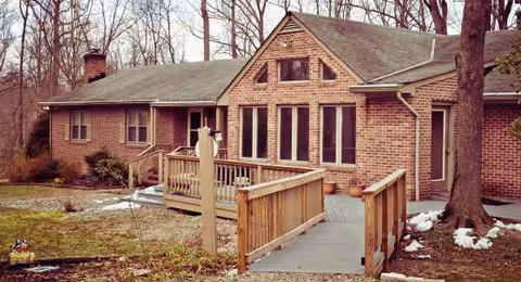 Brick assisted living residence with a wooden ramp leading to the entrance, surrounded by trees.