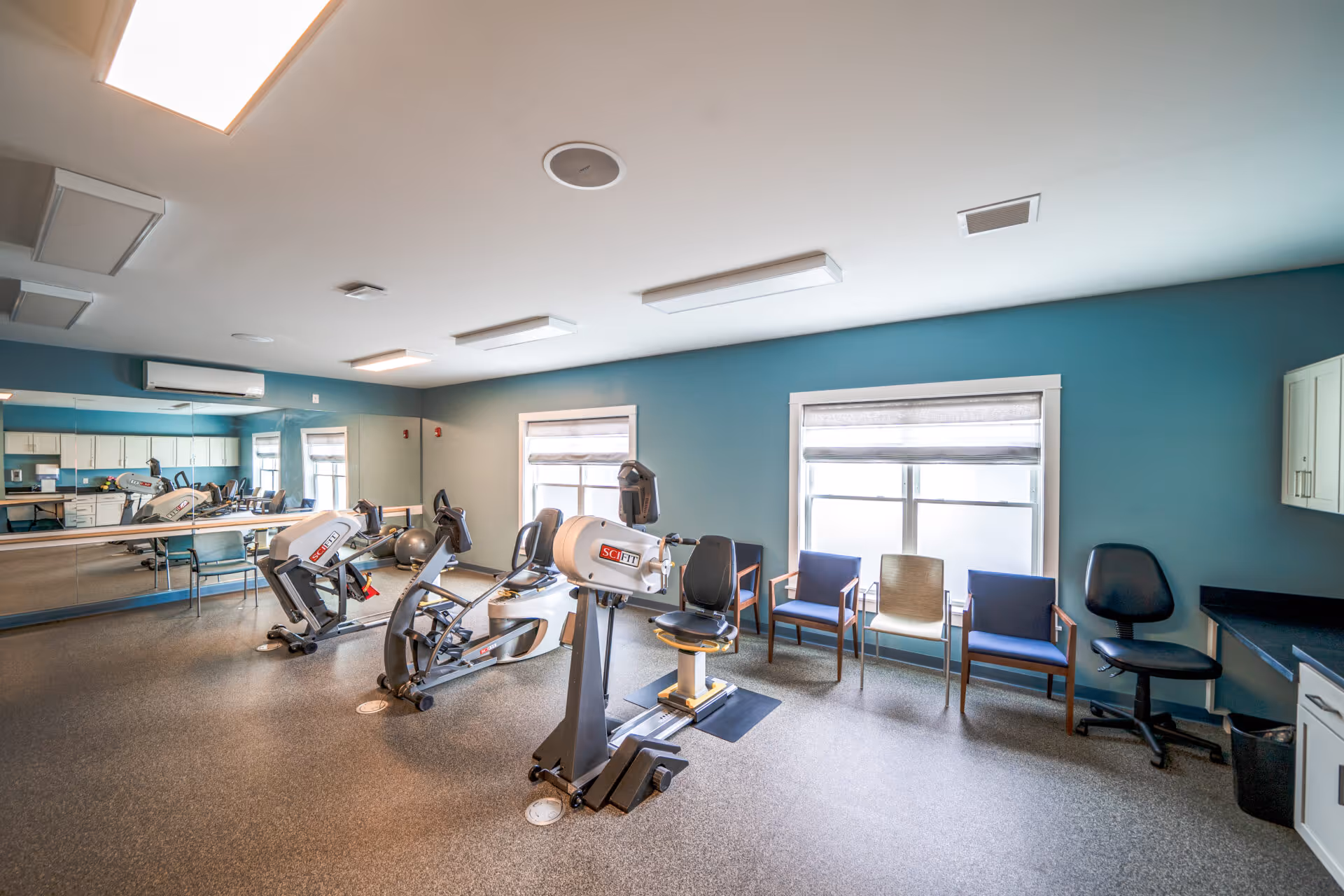 A fitness room with exercise equipment including stationary bikes and a SciFit machine, several chairs lined up against a blue wall, large windows with blinds, a mirrored wall reflecting the room, and overhead fluorescent lighting.