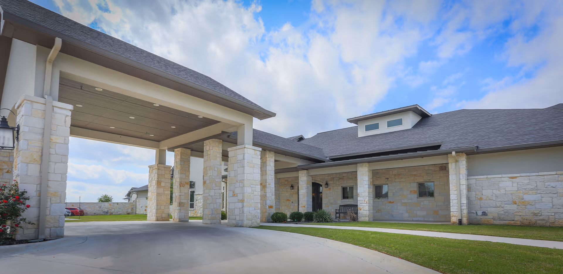 Exterior view of Arbor House Assisted Living and Memory Care building with stone pillars supporting a covered driveway entrance, a well-maintained lawn, and a partly cloudy sky.